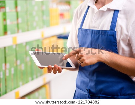 Sales clerk wearing apron using a digital tablet with store shelves on background.