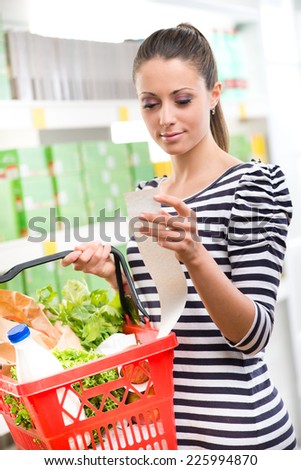 Smiling woman examining a long receipt at supermarket and holding a full shopping basket.