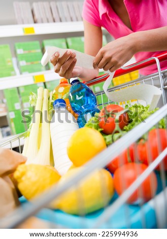 Unrecognizable woman checking a long grocery receipt leaning to a full shopping cart at store.