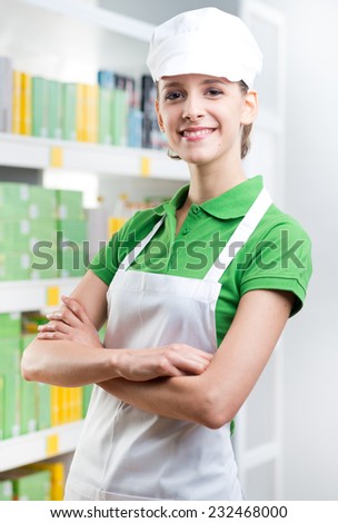 Young female sales clerk with crossed arms smiling at camera with supermarket shelf on background.