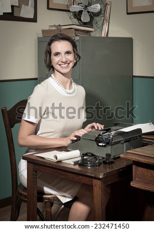 Young smiling secretary at office typing on a vintage typewriter and looking at camera.