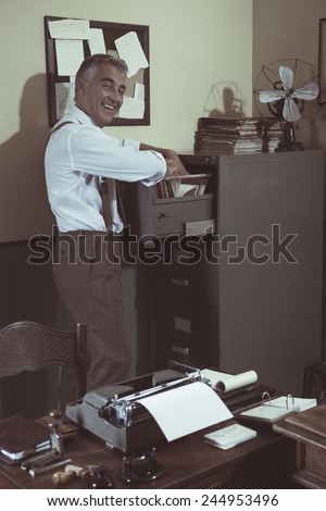 Cheerful smiling employee in 1950s style office searching for a file in the cabinet.