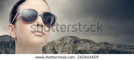 Young female fashion model wearing big sunglasses on mountains and cloudy sky on background