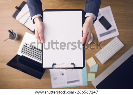 Businessman working at office desk and showing a clipboard with a blank document, computers and stationery on background, top view