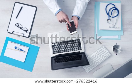 Doctor working at office desk and using a mobile touch screen phone, computer and medical equipment all around, top view