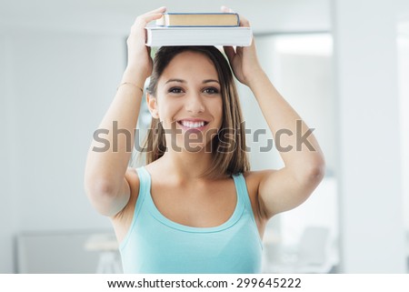 Cute smiling teen student carrying books on her head and looking at camera, education and learning concept