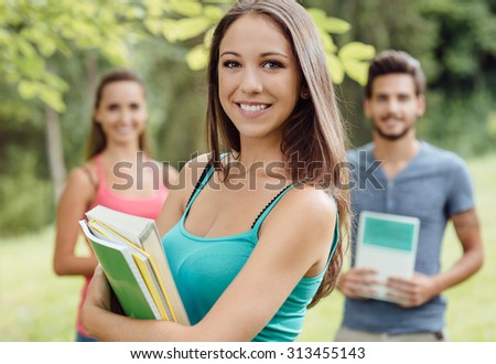 Smiling young student at the park holding books and notebooks, her friends are standing on background