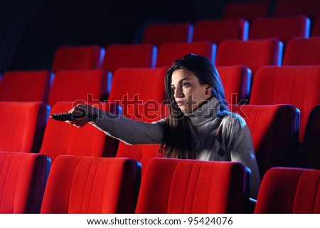 a young woman holding a TV remote control, in the background you can see the red chairs in a movie theater. conceptual image
