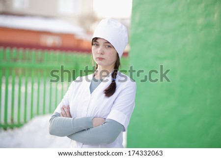 Outdoor portrait of young medical student in white uniform on green fence background.