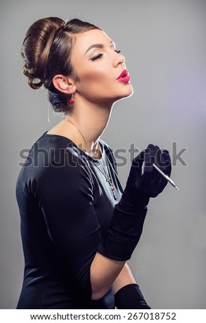 Portrait of young woman in black dress pretending to smoke with a makeup brush on a grey background