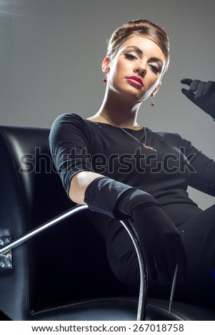 Elegant beautiful woman in a black cocktail dress and long gloves sitting in a black contemporary armchair on a grey background