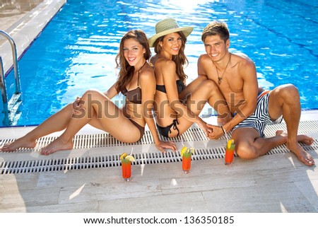 High angle view of two beautiful women in bikinis enjoying drinks at the edge of a sparkling blue swimming pool with a handsome young man