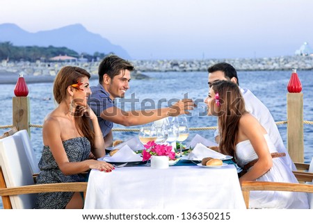 Two couples celebrating at the sea seated at a table at an open-air waterfront restaurant enjoying glasses of cold white wine as they wait for their meal