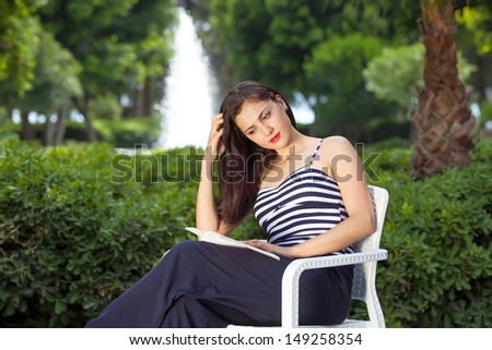 Young beautiful woman reading book in park, sitting outdoors on chair, concentrated on reading book.