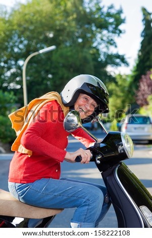 Vivacious elderly woman riding a scooter. Vivacious elderly woman riding a scooter dressed in trendy casual clothes and wearing a safety helmet laughing as she rides by in the street
