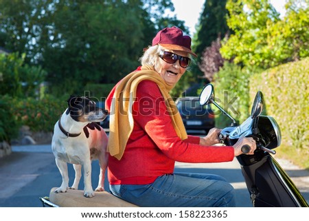Laughing senior woman riding a scooter with her dog. Elderly lady laughing happily as she takes her dog, a small jack russell terrier, for a scooter ride balanced on the pillion behind her