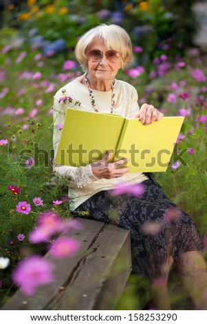 Elderly stylish woman wearing sunglasses sitting in her garden on a wooden bench amongst the flowers with a book enjoying the summer sun