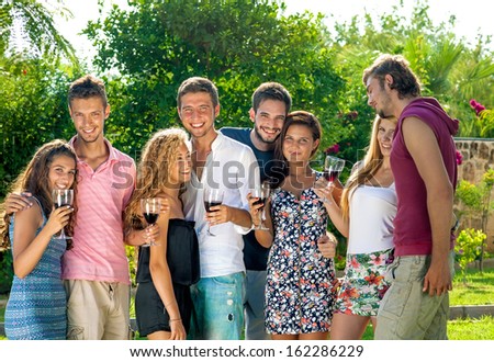 Group of happy young teenage couples standing arm in arm celebrating the summer vacation drinking wine together in the garden laughing and smiling