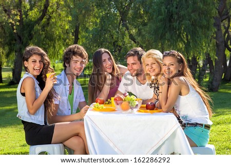 Group of happy vivacious teenagers full of vitality enjoy a fruity meal together sitting around a picnic table outdoors in the park