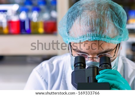 Male chemist working in a laboratory wearing glasses, a surgical cap and mask looking down the ocular of his microscope as he performs an analysis , close up of his head