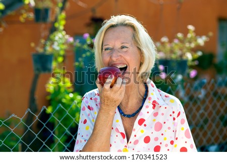 Laughing senior woman eating a red apple opening her mouth wide to take a bite as she stands in the garden in the sunshine.