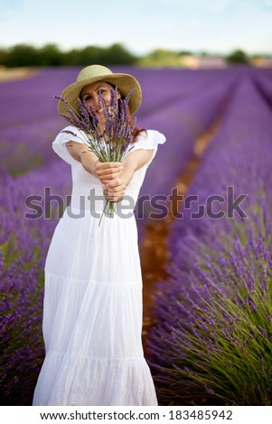 Beautiful female holding a bouquet of lavender twigs in both hands, arms outstretched as she wants to present the bouquet to the viewer. Looking into camera. Sky visible.