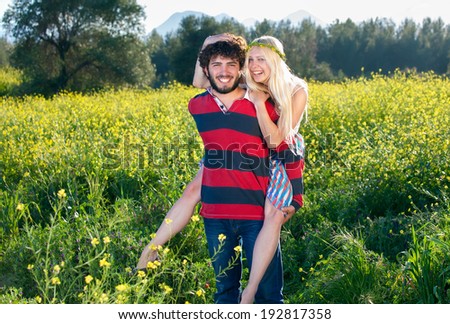 Happy bohemian young man and woman with flowers garlands in the hair laughing as he gives his girlfriend a piggy back ride through a colorful yellow rapeseed field
