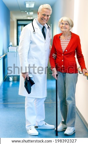 Senior male doctor with an elderly woman patient at the hospital standing together in the passage supporting her by the arm as she walks with a cane  both smiling happily at the camera.