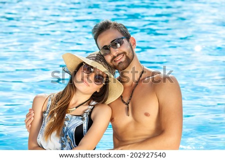 Affectionate happy couple relaxing at a sparkling sunlit blue pool sitting leaning their heads together as they smile at the camera, man is shirtless, woman in sunhat
