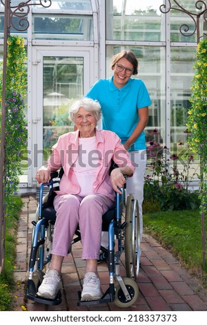 Smiling happy elderly lady in a wheelchair posing with an attractive young care assistant in front of the care home on a bricked pathway
