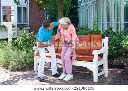 Care assistant tending to a senior lady helping her to make herself comfortable on an outdoor wooden bench in the garden of the care home.