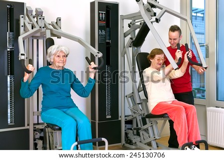 Two Healthy Old Women Exercising Using Chest Press Equipment at the Fitness Gym with Young Male Instructor