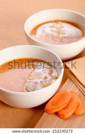 Two bowls of carrot soup with milk foam and sliced almonds. Close to the bowls a napkin on a cutting board and three slices of carrots.