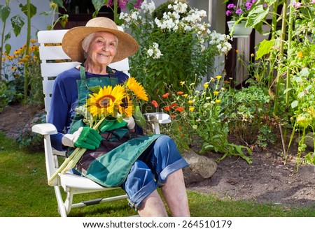 Smiling Old Woman with Hat  Apron and Gloves for Gardening Sitting on a White Chair Holding a Bouquet of Sunflowers  Looking at the Camera.