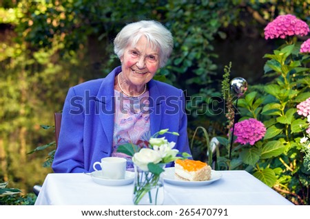 Smiling Old Woman in Blue Violet Blazer Sitting at the Garden Table with Coffee and Cake, Smiling at the Camera.