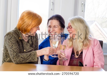Three Happy Middle Age Mom Best Friends Tossing Glasses of White Wine and Smiling Each Other While Sitting at the Wooden Table Inside a Restaurant.