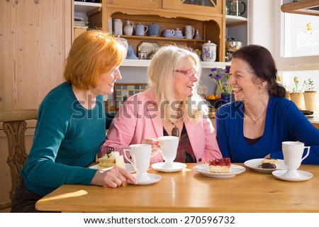 Three Happy Adult Female Friends Having Cups of Tea or Coffee and Slices of Cakes at the Wooden Table While Talking Funny Moments.