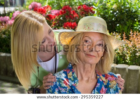Portrait of Young Blond Nurse Peeking Around Shoulder of Senior Blond Woman Wearing Sun Hat Outdoors in Flower Garden of Retirement Building.
