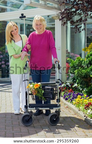 Blond Smiling Nurse Holding Arm of Senior Woman with Walker  Helping Senior Resident to Walk Outdoors in front of Building Entrance Near Flower Beds.