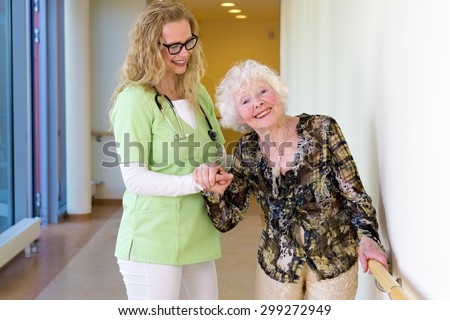 Kind Young Medical Assistant Assisting a Happy Elderly Woman Patient Walking at the Corridor in the Hospital.