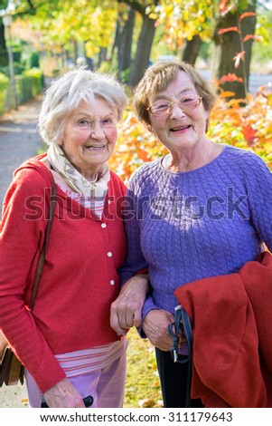 Three Quarter Shot of Two Smiling Senior Women in Autumn Outfits Against Tall Trees at the Street Side.