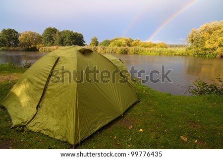green tent pitched on the bank of a lake, with the sky, rainbow, trees, lake visible in the foreground
