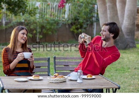 Smiling man photographing his girlfriend or wife while enjoying coffee and cakes at an outdoor cafe