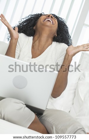 African American woman at home sitting on sofa or settee celebrating and laughing using her laptop computer.