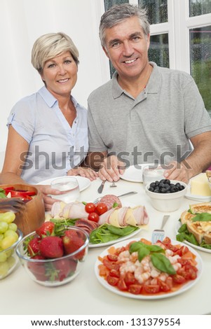 An attractive happy, smiling middle aged couple healthy eating salad, seafood, cold meats and fruit at a dining table.