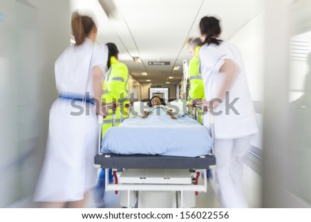 A motion blurred photograph of a young Asian Indian girl child patient on stretcher or gurney being pushed at speed through a hospital corridor by doctors & nurses to an emergency room