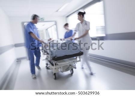 A motion blurred photograph of a senior female patient on stretcher or gurney being pushed at speed through a hospital corridor by doctors & nurses to an emergency room Stock fotó © 
