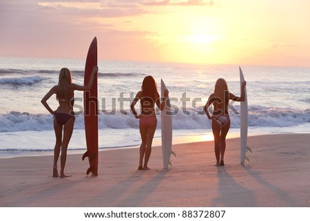 Rear view of three beautiful young women surfer girls in bikinis with surfboards at a beach at sunset or sunrise