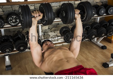 Bodybuilder working out in a gym lying on his back lifting weights against a backdrop of shelving loaded with exercise equipment