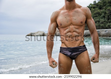 Strong muscular fit man posing in a swimsuit on a tropical beach showing off his powerful physique, anonymous torso view